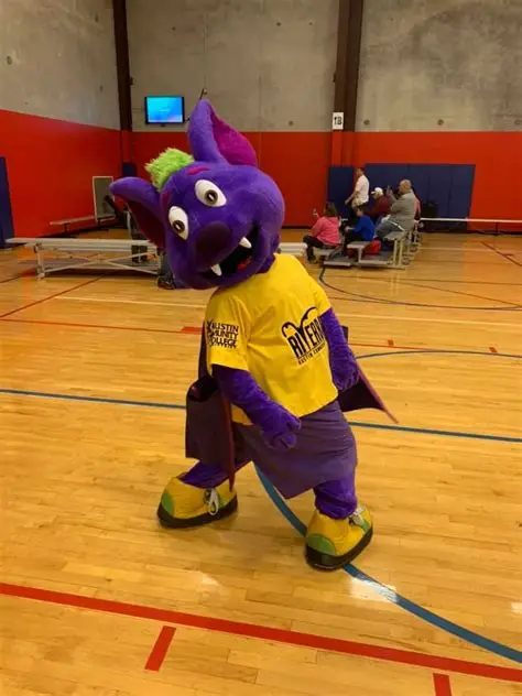 Purple bat mascot wearing a yellow Austin Community College shirt stands on a basketball court.